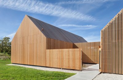 Wood-clad modern building with vertical slats and a sloped roof, set on a grassy site with a concrete path.